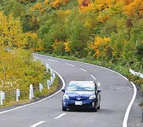 Blue sedan driving on a winding road surrounded by autumn foliage, illustrating car rental services in Jaipur